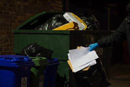 Gloved hand pulling sensitive paper documents from an overflowing dumpster at night, illustrating the risk of identity theft and data breaches from improper document disposal.